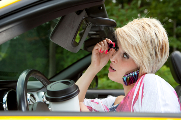 Distracted driver applying mascara while talking on the phone and holding coffee in a car, demonstrating dangerous driving behavior.