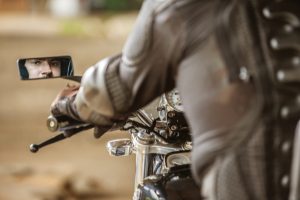 Motorcyclist in protective gear looking into rearview mirror with focused expression on a classic motorcycle.