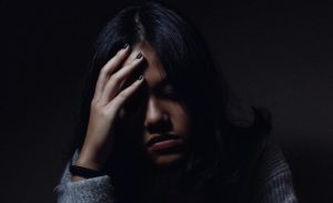 Young woman in low light holding her head in distress, representing anxiety, depression, or emotional struggle.