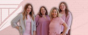Amanda Hall team of four women standing together and smiling in front of a pink brick wall with a feather and column logo; they are dressed in shades of pink and light colors.