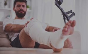 Injured man with bandaged leg sitting on couch next to crutches, recovering from accident or personal injury.