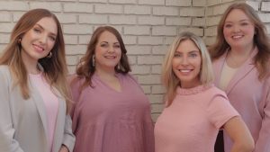 Amanda Hall Law Team — four women standing in front of a white brick wall, smiling and dressed in pink and light-colored outfits.
