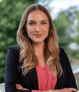 Amanda Hall Law - Lawyer Tessa Martin Woman with long, wavy blonde hair wearing a dark blazer and pink blouse, standing outdoors with green trees in the background, smiling confidently with arms crossed.