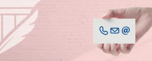 A hand holds a white card displaying blue icons for phone, email, and online contact against a pink brick wall with a stylized feather and column logo, symbolizing communication and professional outreach.