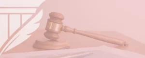 Wooden gavel resting on a sound block beside legal papers and a pen, with a feather-shield emblem in the pink-tinted background, symbolizing justice, legal authority, and institutional branding.