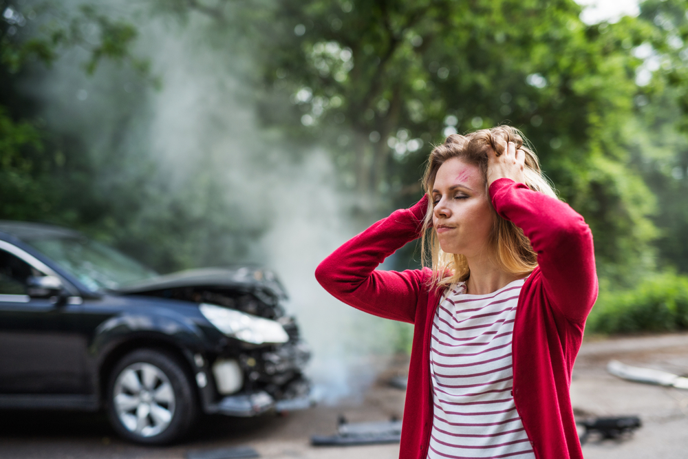 Woman holding her head after a car accident, standing near a damaged vehicle with smoke in the background.