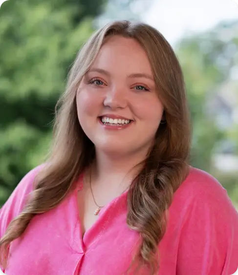 Amanda Hall Law Firm Paralegal-Chloe Barner. Woman with long, wavy light brown hair wearing a bright pink blouse, smiling warmly while standing outdoors with greenery in the background.