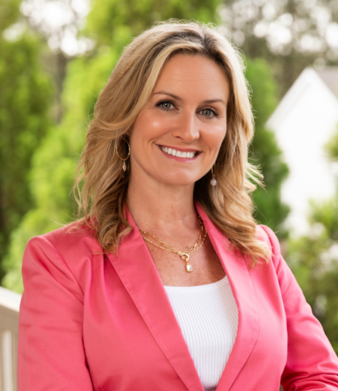 Portrait of a smiling woman with blonde hair wearing a pink blazer and white top, standing outdoors with greenery in the background.