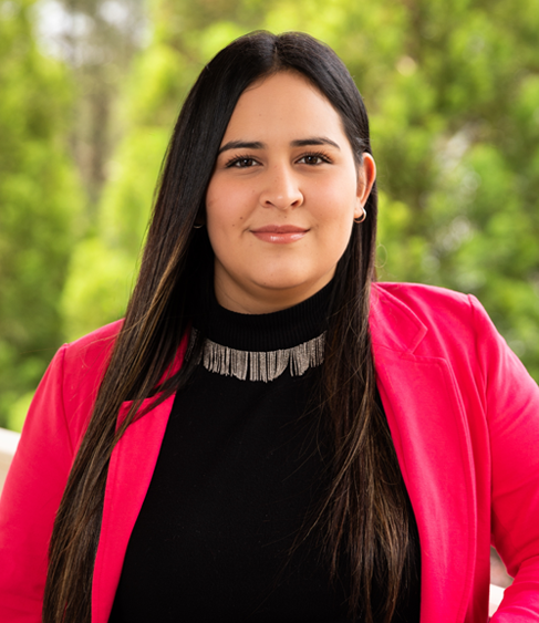 Woman with long dark hair wearing a bright pink blazer and black turtleneck, outdoors with a green blurred background.