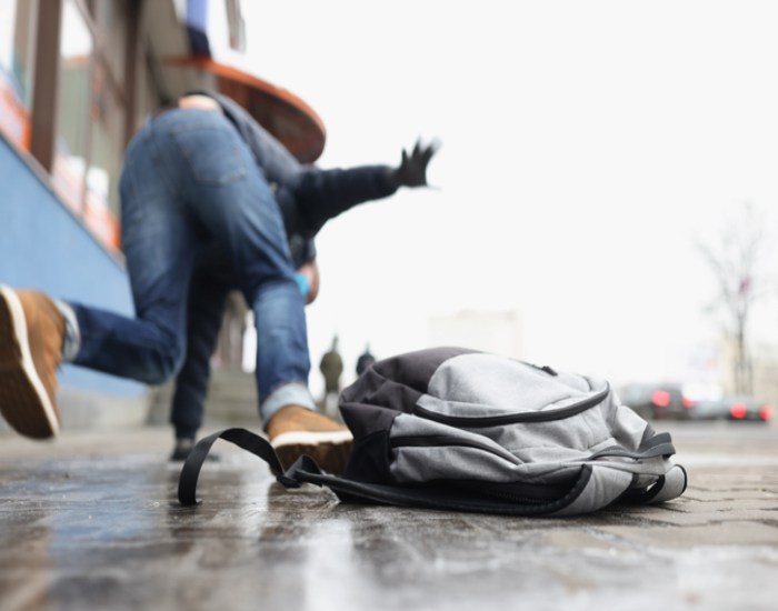 Gray backpack lying on a wet sidewalk as a person in jeans and a hat stumbles in the background near a storefront.